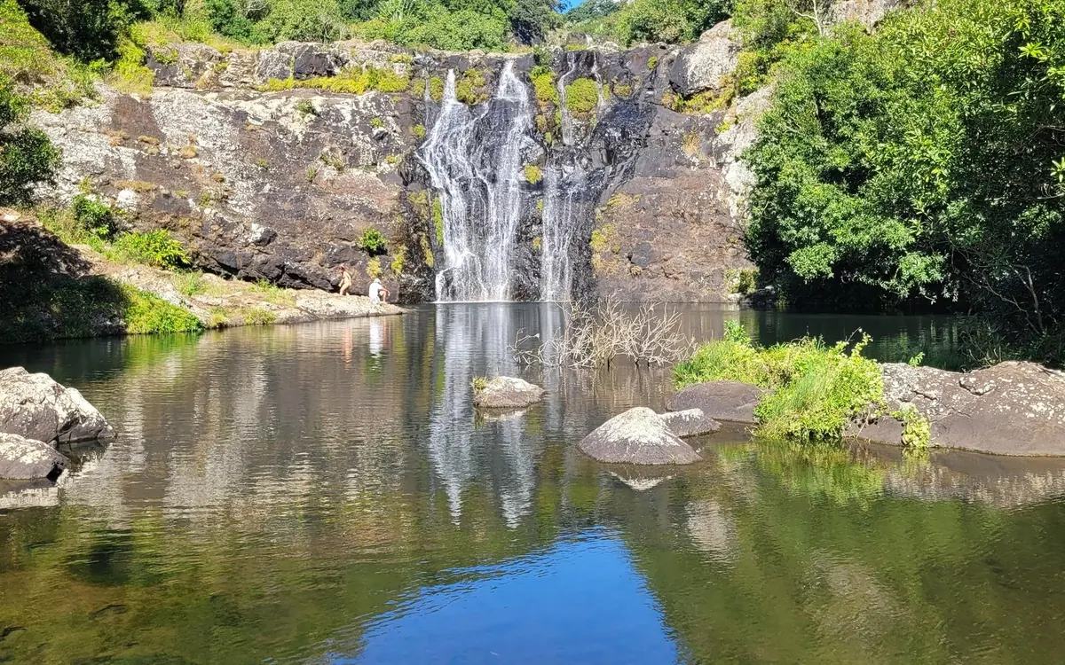 A wide pool below a dark basalt wall on the island, with water running down the rock face and two people standing at the edge