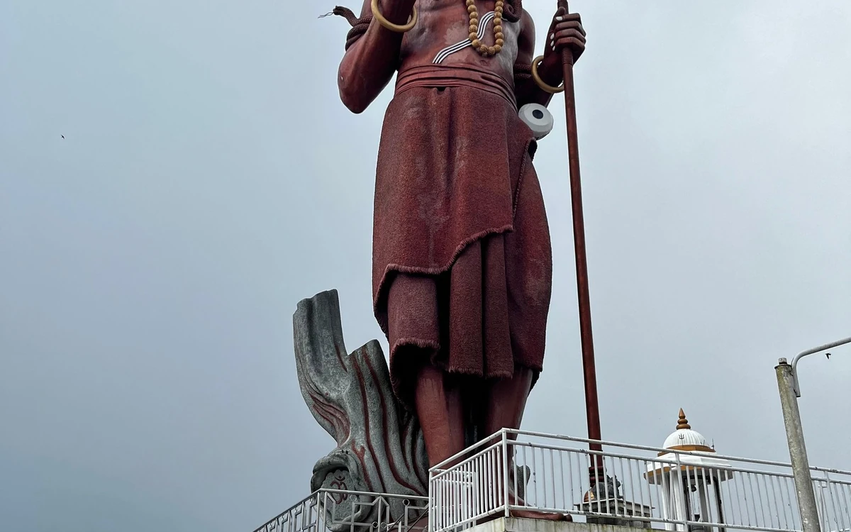 A tall statue seen from below against a grey sky, with a railing and a temple roof beside it